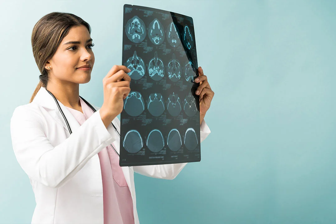 A young female doctor in a white coat carefully examining an X-ray film with medical scans visible, in a bright clinical setting with light blue walls
